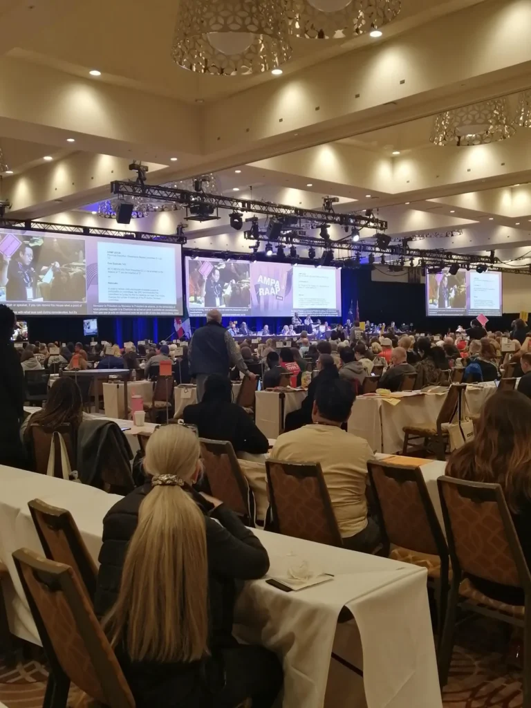 A large conference hall filled with people seated at tables facing a stage with multiple screens displaying presentations and speakers. The room has high ceilings and decorative lighting fixtures.