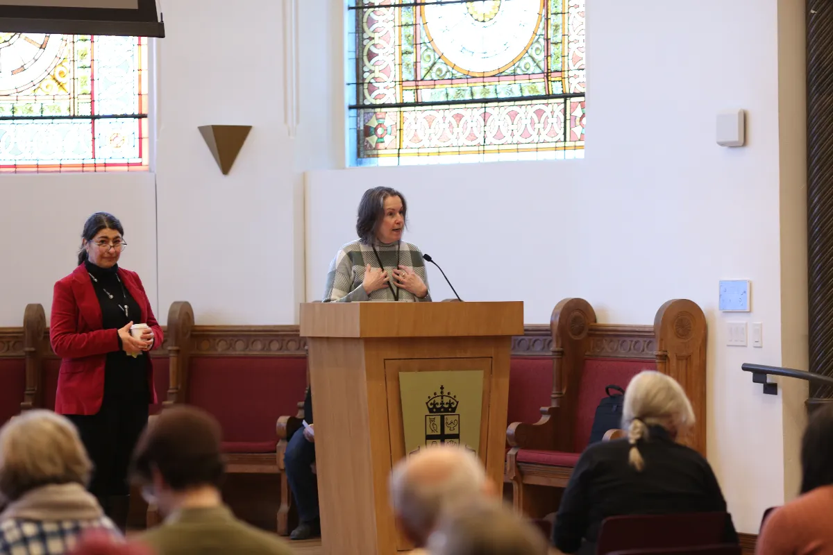 A woman stands at a podium speaking to an audience in a room with stained glass windows; another woman in a red blazer stands nearby, holding something in her hands.