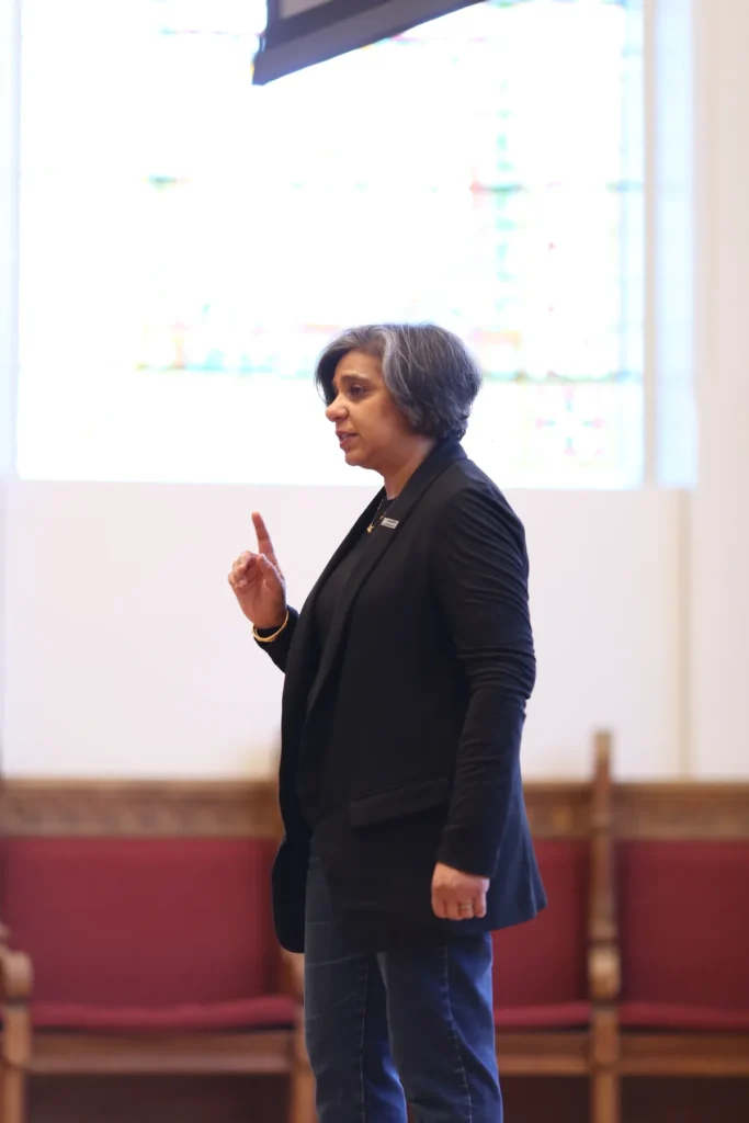 A person with short gray and black hair, wearing a dark blazer and jeans, stands indoors with one finger raised, speaking. Red cushioned chairs and a bright window are in the background.