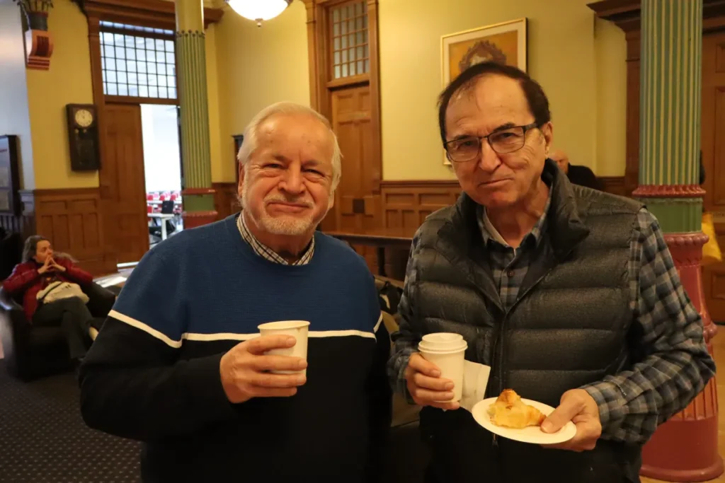 Two older men stand indoors, smiling at the camera. One holds a cup, the other holds a cup and a plate with a pastry. A woman sits in the background on a couch. The setting appears to be a cozy, well-lit room.