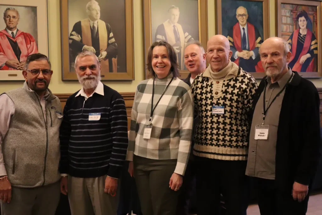 Six adults stand in a row indoors, smiling at the camera. Behind them are framed portraits of people in academic robes on the wall. Four men have beards, and one woman stands in the center, all wearing name tags and winter clothing.