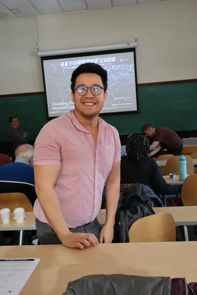 A smiling person in glasses and a striped shirt stands at the front of a classroom with seated people and a presentation slide titled BEYOND THE CODE displayed on a screen behind them.