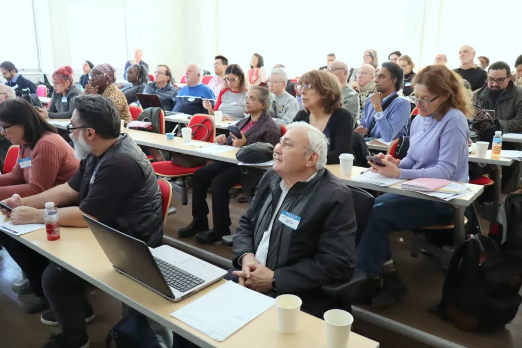 A diverse group of adults sits in a classroom, listening attentively. Some take notes, use laptops or phones, and there are papers, notebooks, and cups on the desks. The atmosphere is focused and engaged.