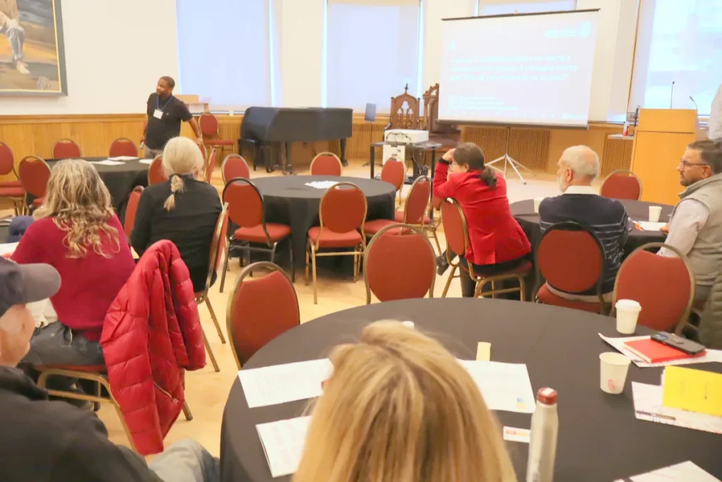 A group of people sit at round tables, listening to a speaker standing at the front of a conference room with a projector screen displaying text in the background.