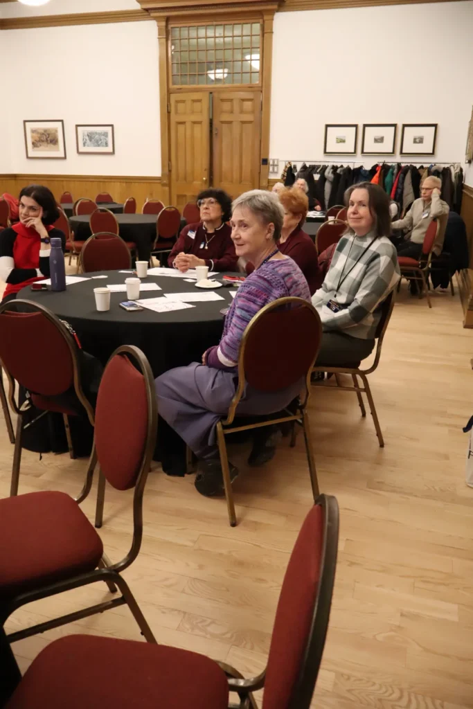 People sit around a round table in a wood-paneled room, looking toward the front. Papers, cups, and a water bottle are on the table. Coats hang on a rack behind them, and framed pictures decorate the walls.