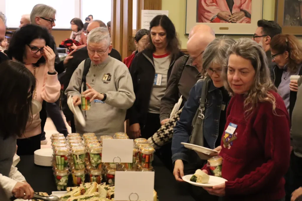 A group of people at a buffet table serve themselves food during an indoor social event. Some hold plates and cups while selecting items, and there are jars of vegetables and sandwiches on the table.