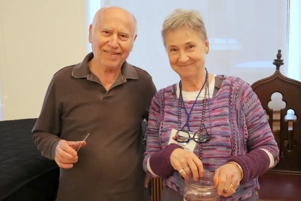 An older man and woman stand side by side indoors, smiling at the camera. The man holds a small tool, and the woman has her hands on a clear container. Both appear relaxed and happy.