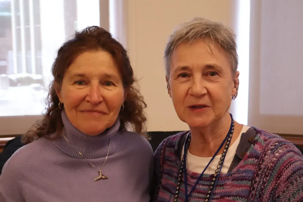Two older women smiling and posing together indoors, both wearing purple sweaters and necklaces, with large windows and light streaming in the background.