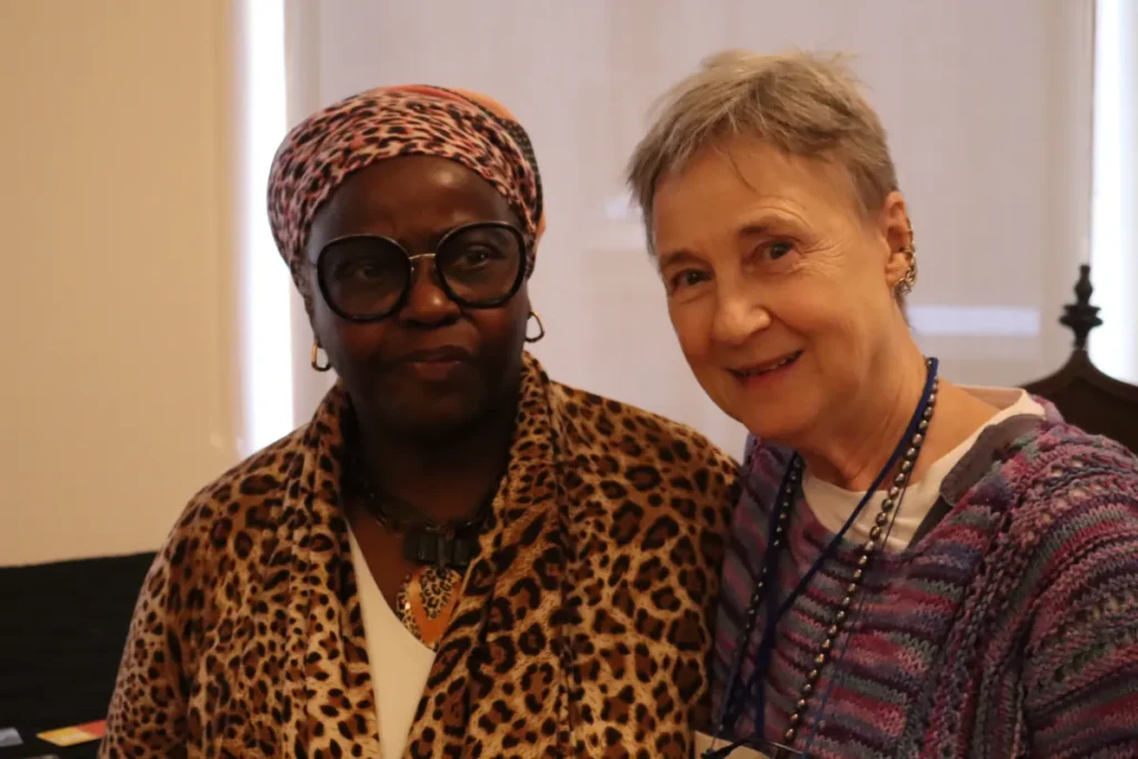 Two women standing close together and smiling. One wears glasses, a leopard print headscarf, and matching jacket; the other has short gray hair and a purple sweater. They appear to be in a warmly lit indoor setting.
