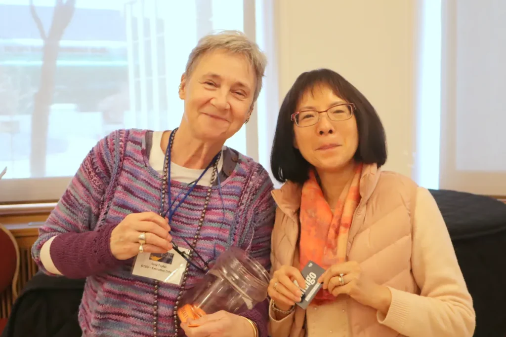 Two women stand side by side indoors, smiling at the camera. One wears a purple-striped sweater and holds a clear jar, while the other wears glasses, a pink vest, and an orange scarf, holding a gift card.