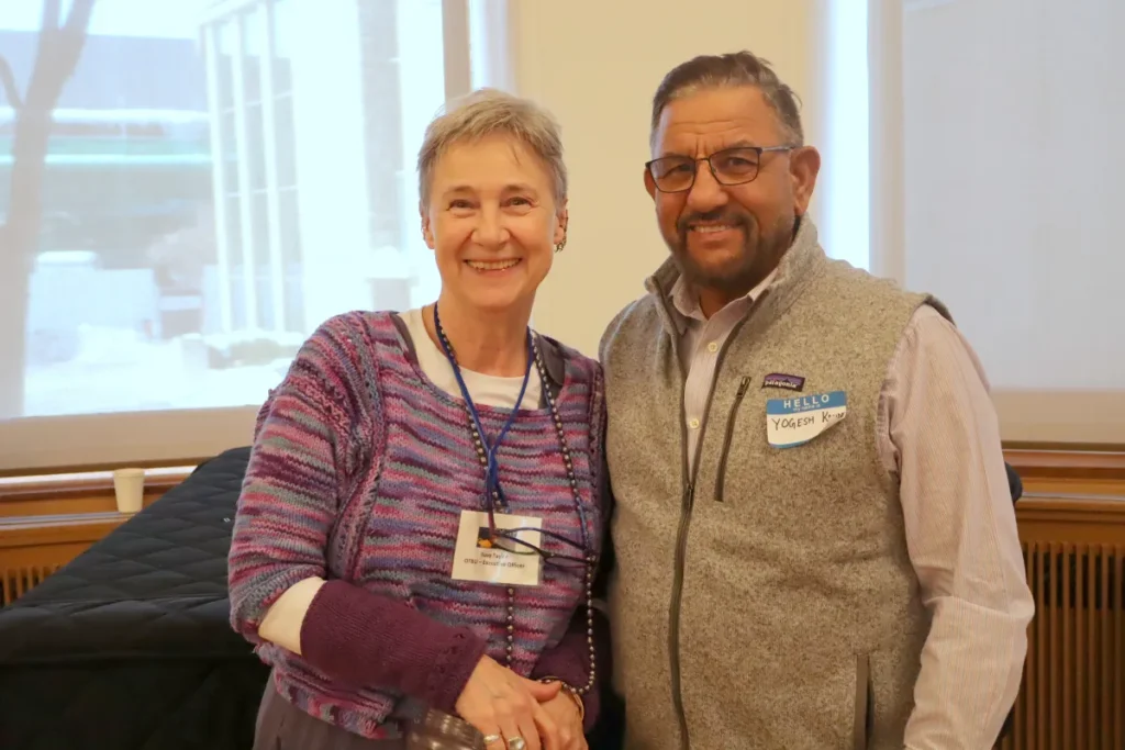 Two adults stand closely together indoors, smiling at the camera. The woman wears a purple sweater and the man wears glasses, a gray vest, and a Hello, My Name Is sticker. A bright window is in the background.