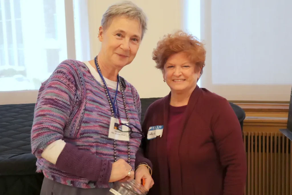 Two older women stand together indoors, smiling at the camera. One woman wears a purple sweater and holds a plastic container, while the other wears a maroon cardigan. Both have name tags and short hairstyles.