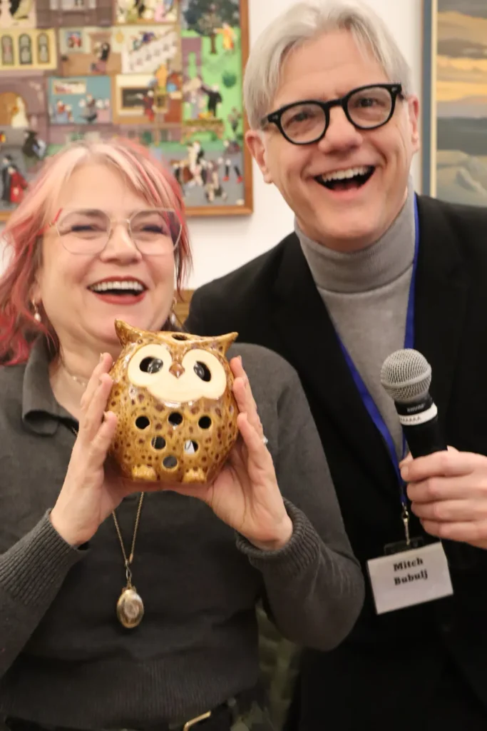 Two smiling adults pose indoors; the woman holds a decorative ceramic owl and wears glasses, while the man, also in glasses and holding a microphone, wears a name tag that reads Mitch Dubuij. Colorful paintings hang behind them.