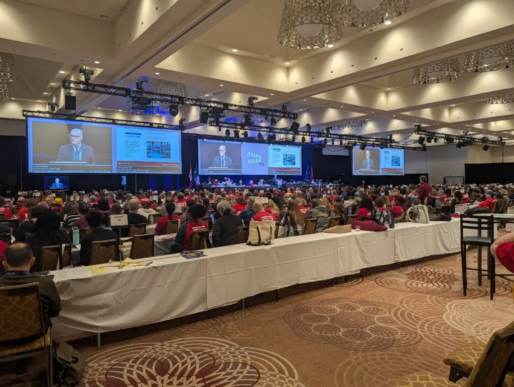 A large conference hall filled with people seated at tables, many wearing red shirts. Several large screens display a speaker at a podium addressing the audience. The room is well-lit with modern chandeliers.