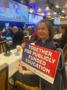 A woman sits at a conference table holding a sign that reads, “TOGETHER FOR PUBLICLY FUNDED EDUCATION.” Other attendees and a large screen are visible in the background.