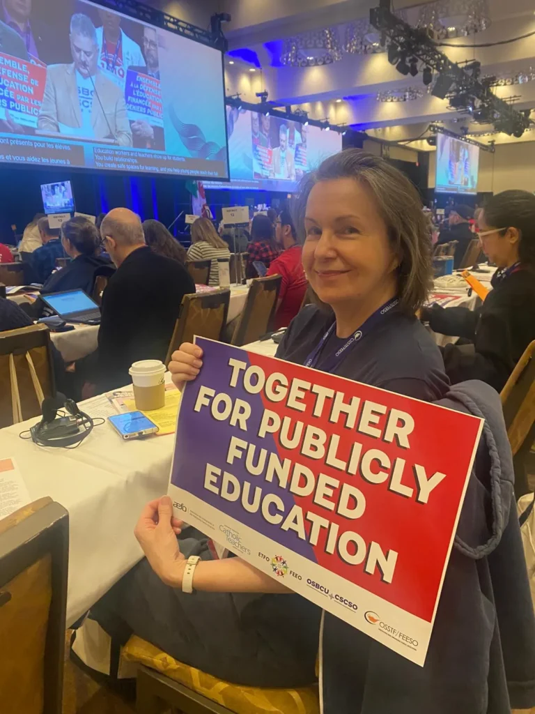 A woman sits at a conference table holding a sign that reads, “TOGETHER FOR PUBLICLY FUNDED EDUCATION.” Other attendees and a large screen are visible in the background.