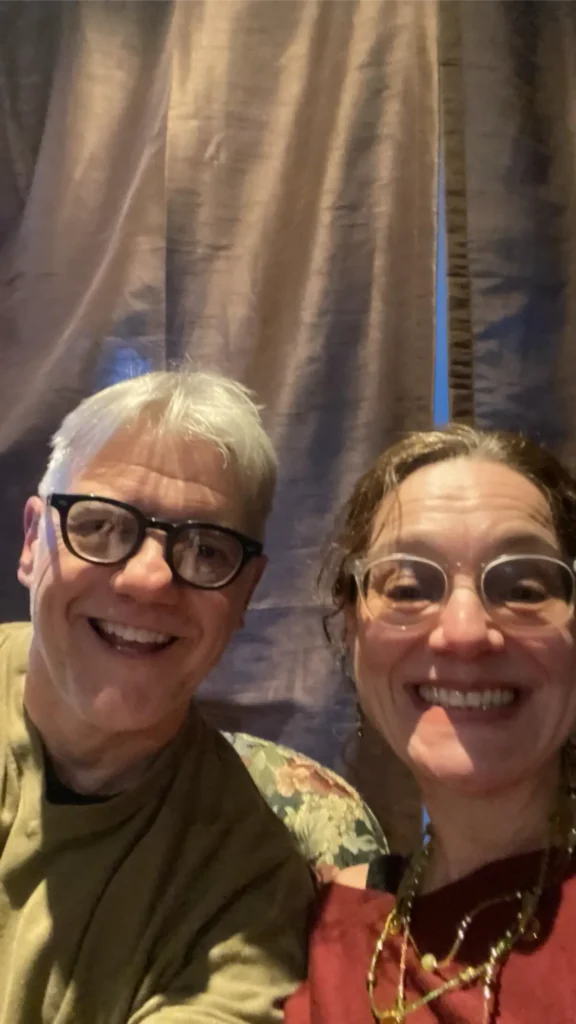 Two smiling adults wearing glasses pose for a selfie in front of dark curtains. The lighting is soft and warm, and both appear happy and relaxed.