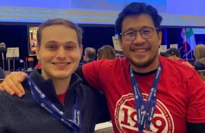 Two people at an event smile at the camera, both wearing blue lanyards; one wears a red t-shirt with “1919” on it, the other a gray shirt. Crowds and event signs are visible in the background.